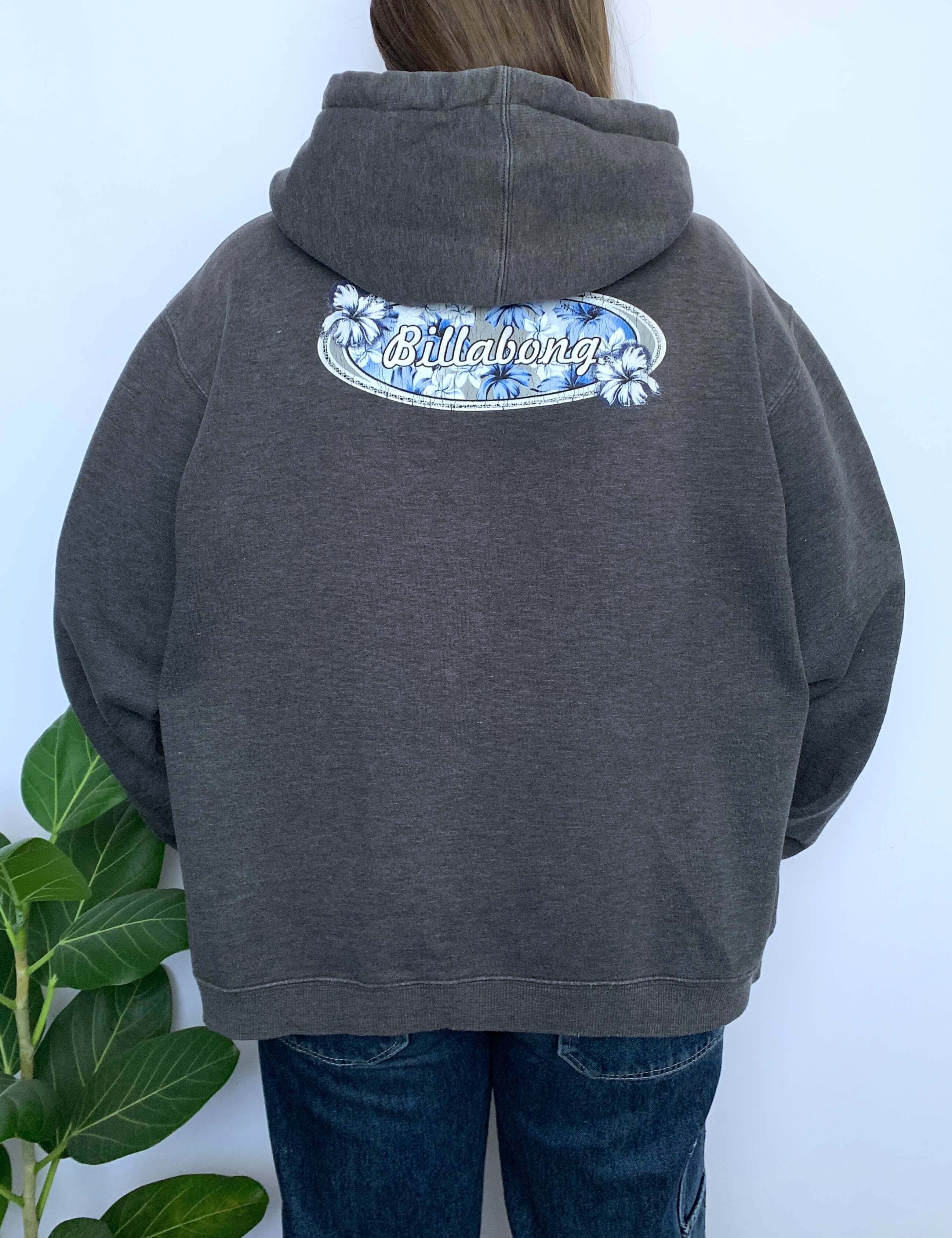 Person wearing a grey hoodie with 'Billabong' logo on the back against a white wall.