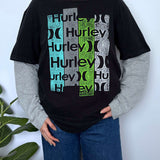 A person wearing a black graphic T-shirt with layered gray long sleeves and blue jeans, standing against a light wall with a plant at the side. The shirt features repeated 'Hurley' text with moon-phase graphics in teal, gray, and green.